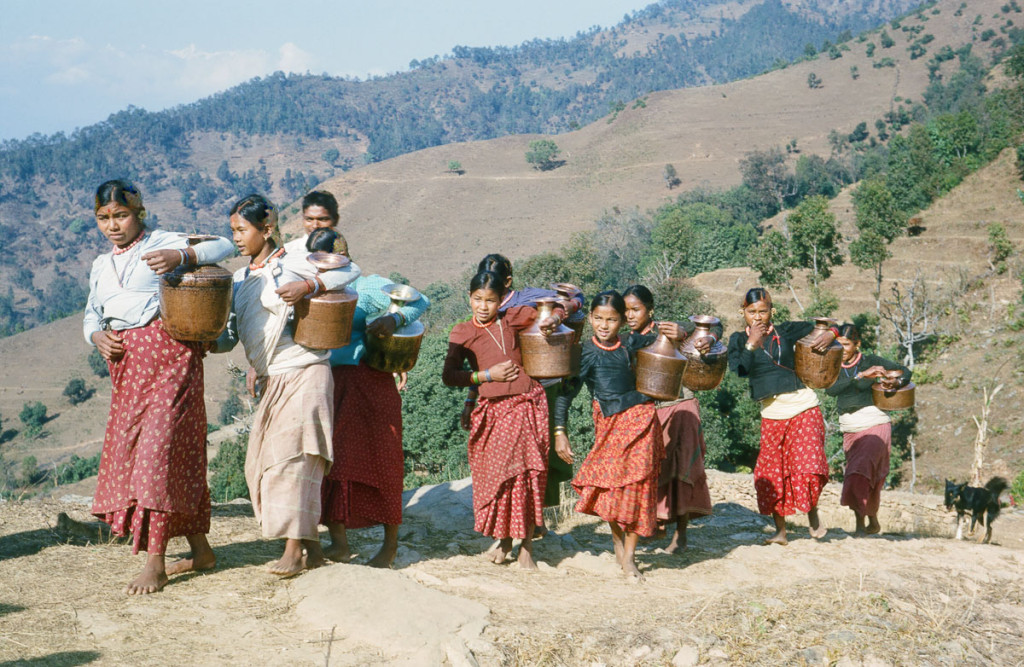 Date: 1964-03 Location: Majhthana, Kaski Description: Brahmin (Bahun) women bringing water up to their village in the dry season. The water source was farther from the village at that time of year, down closer to the Madi Khola. The trail leveled out here and they began singing folk songs.